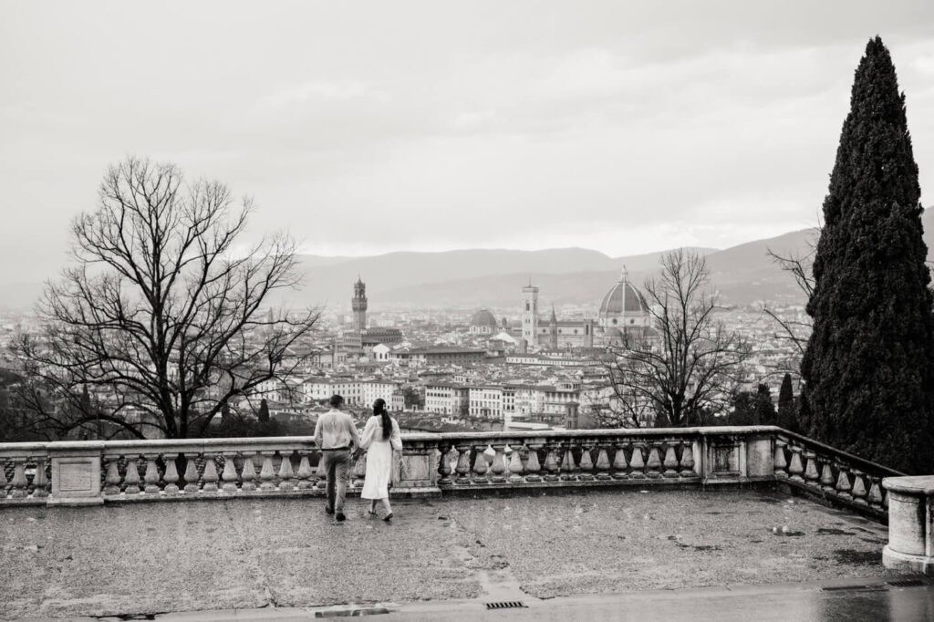 Dreamy walk along the Florence overlook – The couple walks hand in hand along a historic stone terrace, with the Florence skyline stretching behind them.