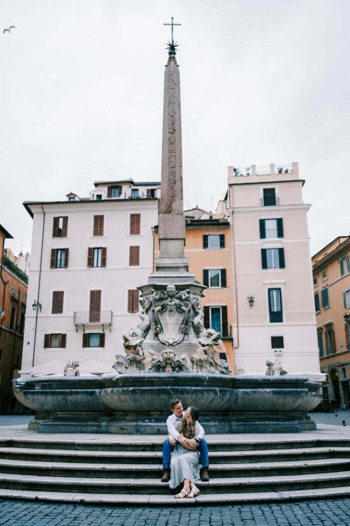 Couple at Fontana del Pantheon – The couple sits on the steps of the Fontana del Pantheon, wrapped in each other’s arms, as the historic fountain and surrounding Roman buildings frame the scene.