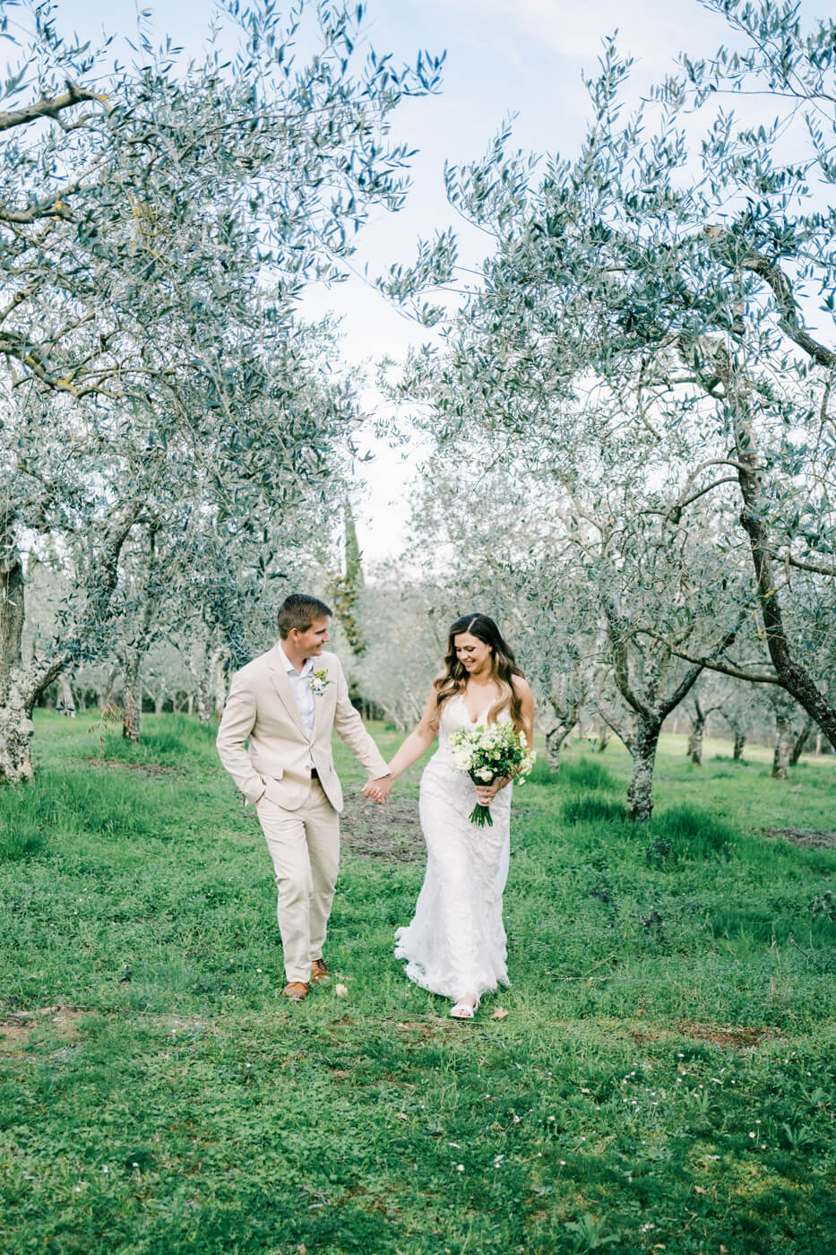 Bride and groom walk hand in hand through an olive grove at Villa Olimpia, Tuscany, during their romantic destination wedding in Italy.