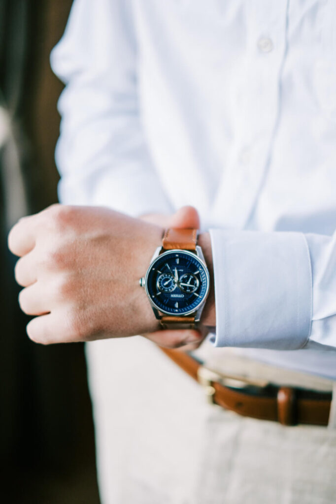 Close-up of groom adjusting his classic blue-faced wristwatch with a brown leather strap before the wedding ceremony.