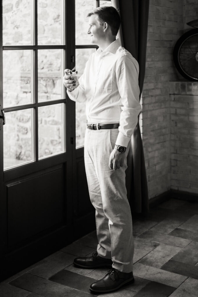 Groom standing by a sunlit window, holding cologne, preparing for his Tuscany villa wedding.