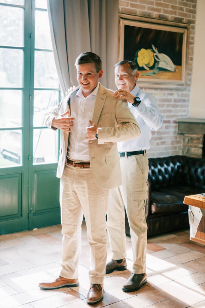 Father helping groom into a tailored beige linen suit, a joyful moment in a Tuscany villa wedding preparation.