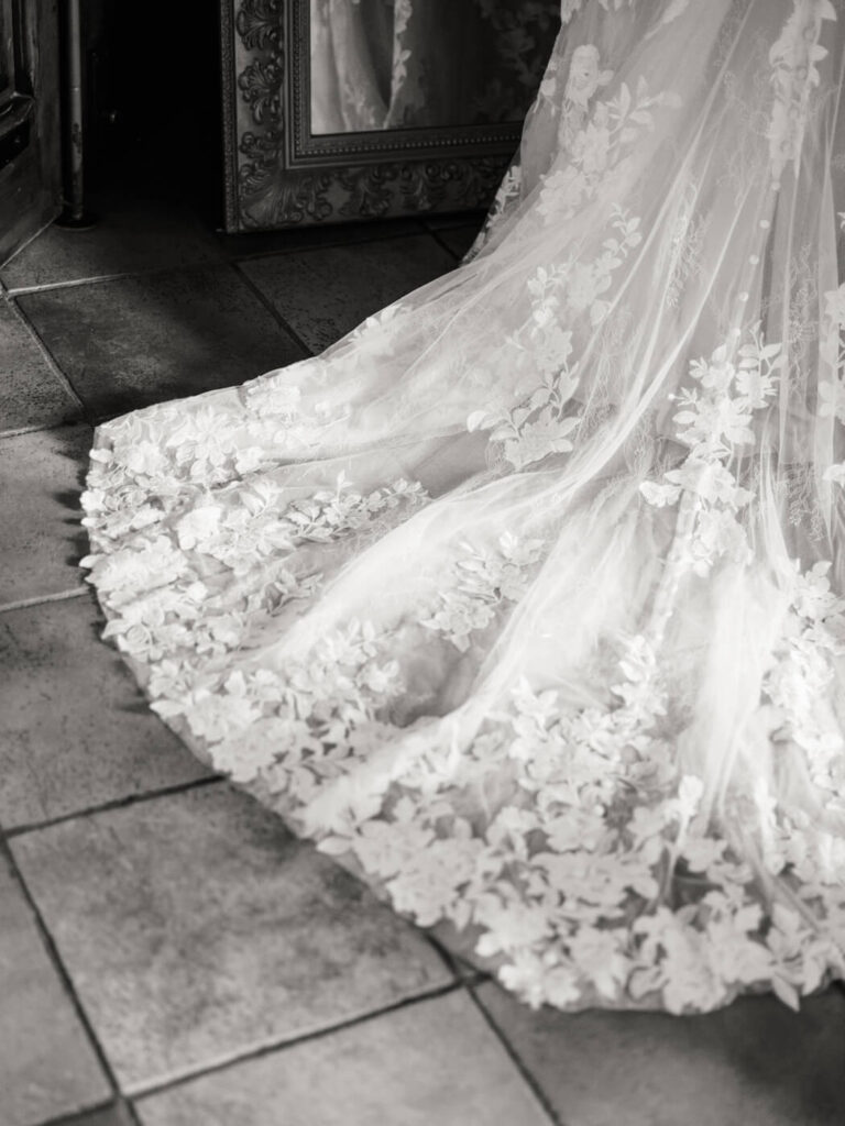 Detailed shot of the bride’s flowing lace wedding gown train, softly resting on rustic terracotta tiles.