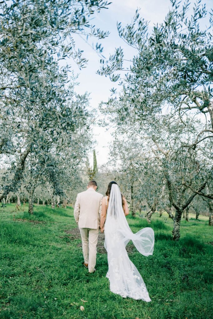 Bride and groom walking hand in hand through a sunlit Tuscany villa courtyard, surrounded by timeless architecture.