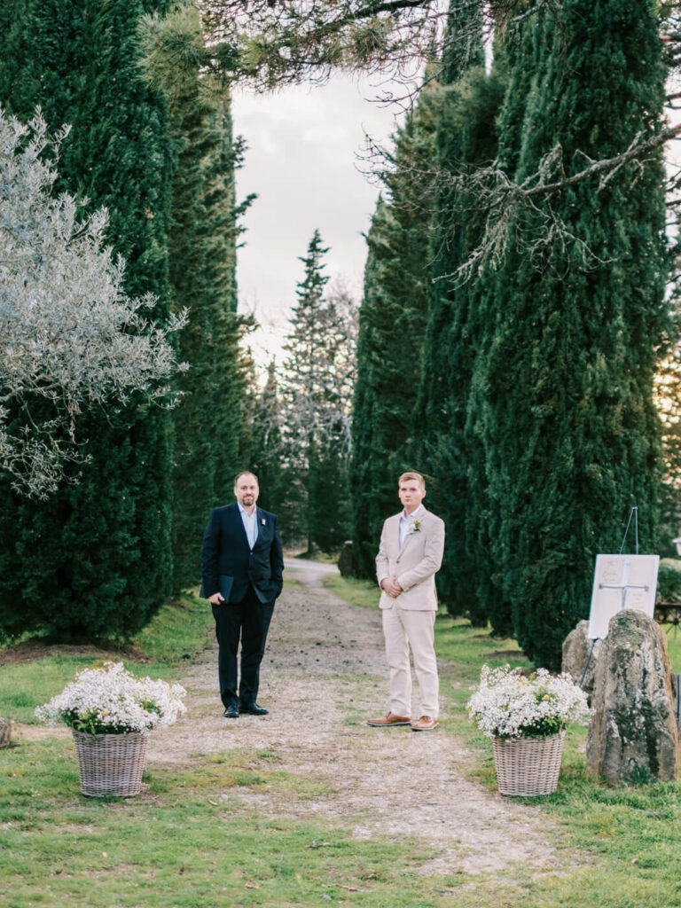 Groom standing with the officiant under the cypress trees, waiting for his bride at a romantic outdoor wedding ceremony.