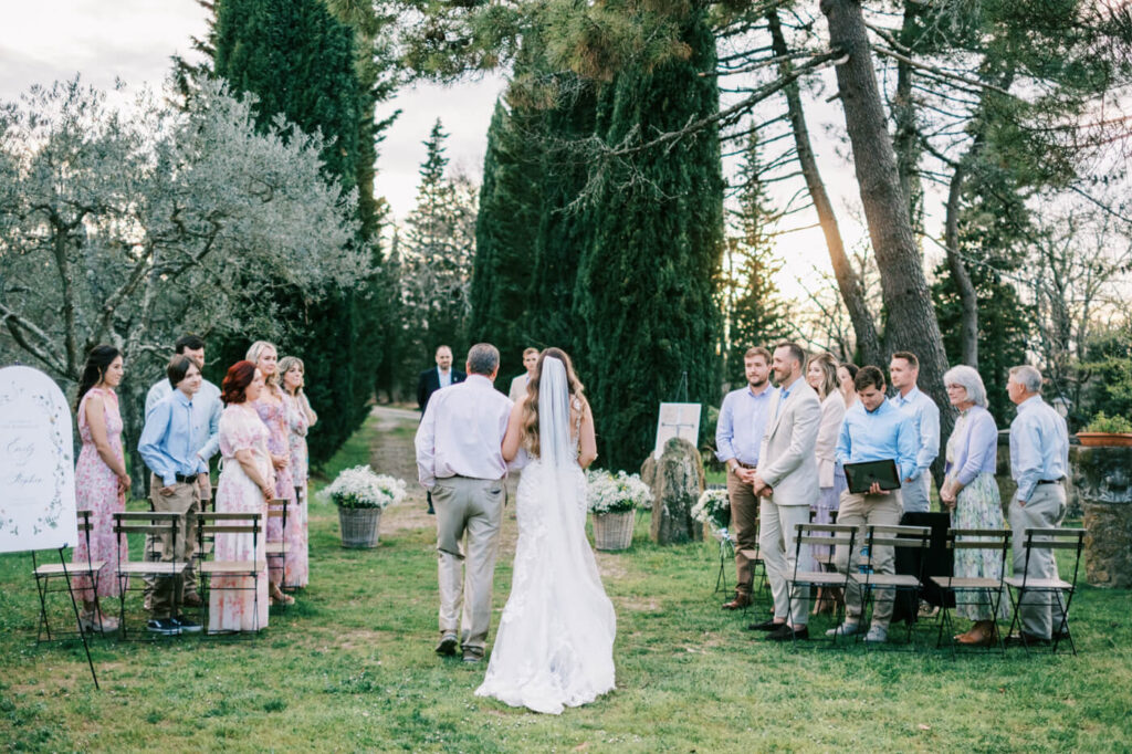 Bride walking down the aisle with her father, surrounded by loved ones and framed by towering cypress trees at golden hour.