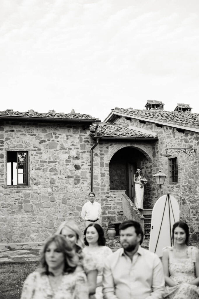 Bride standing on the villa steps, framed by its historic stone walls, preparing for her entrance as guests look on.