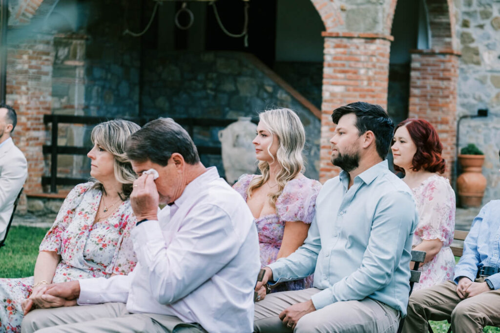 Father of the bride dabs his eyes, overwhelmed with emotion, as family members sit nearby in soft floral pastels.