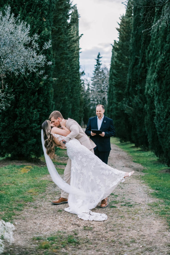 Sealed with a kiss – The groom dips his bride for a romantic first kiss as husband and wife, framed by towering evergreens and the soft light of dusk.