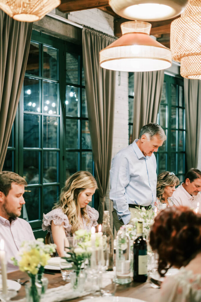 A heartfelt blessing – The father of the groom stands at the head of the table, delivering a touching toast as guests bow their heads in gratitude.