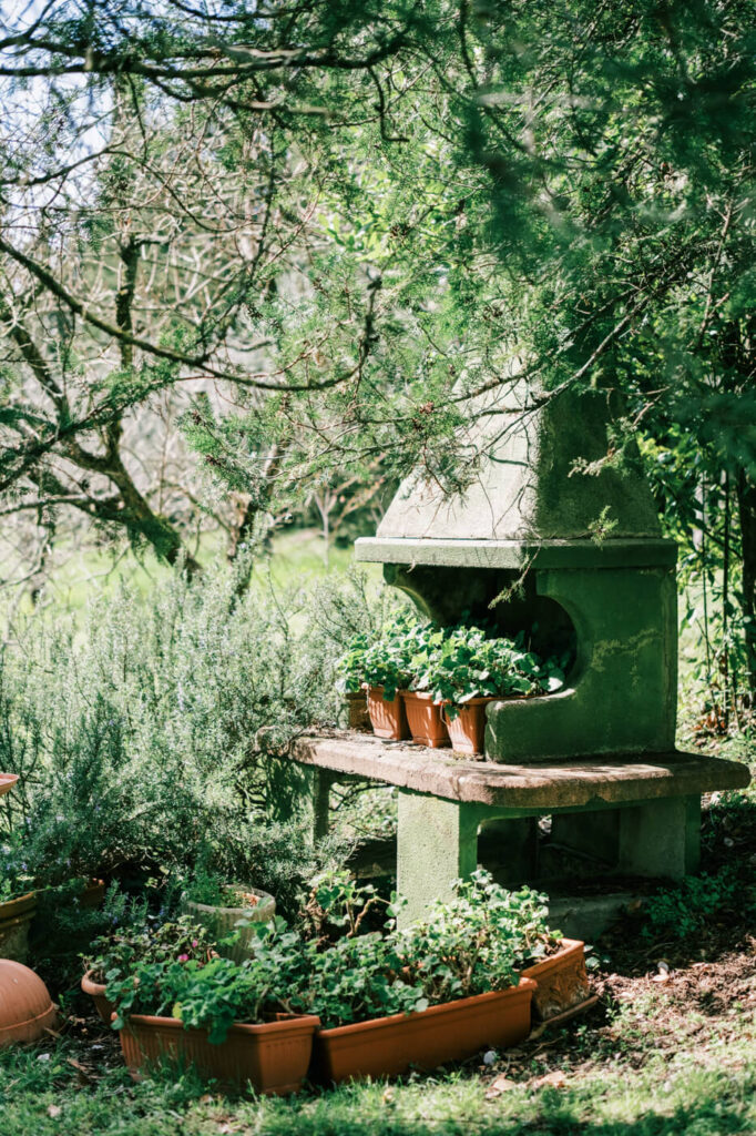 A charming garden space at Villa Olimpia in Tuscany, featuring a rustic green outdoor fireplace surrounded by lush greenery and potted plants.