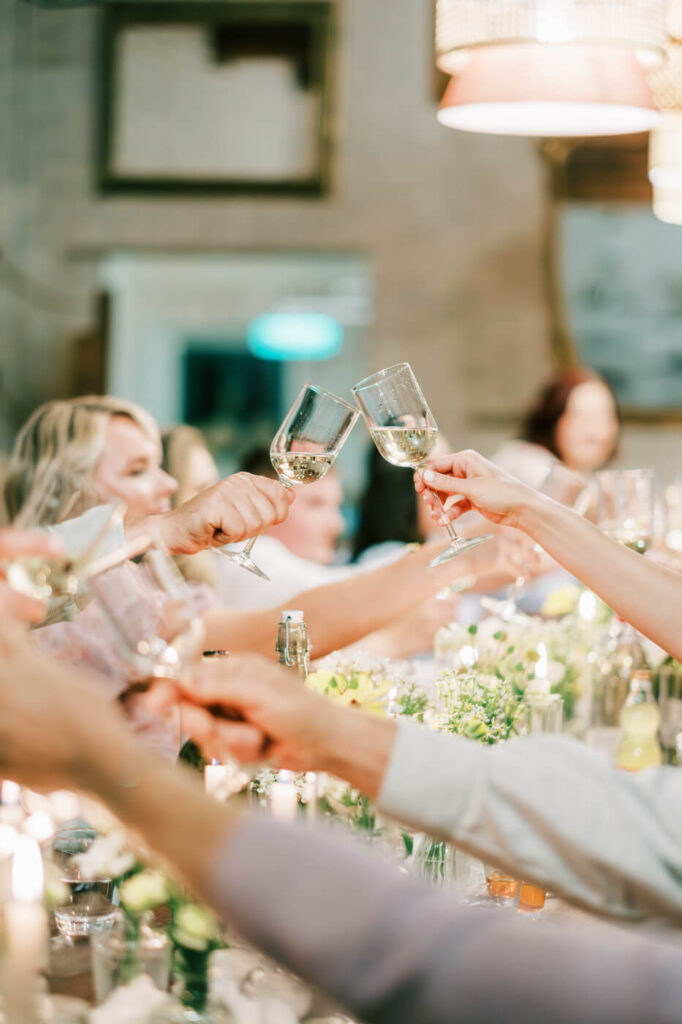 A close-up of wedding guests raising their glasses for a celebratory toast under warm, ambient lighting.