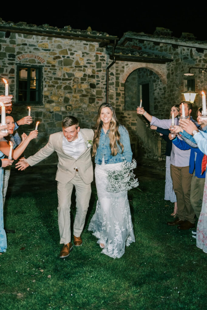 The couple walks hand-in-hand through a candlelit send-off, surrounded by guests holding glowing candles against the backdrop of a Tuscan stone villa.