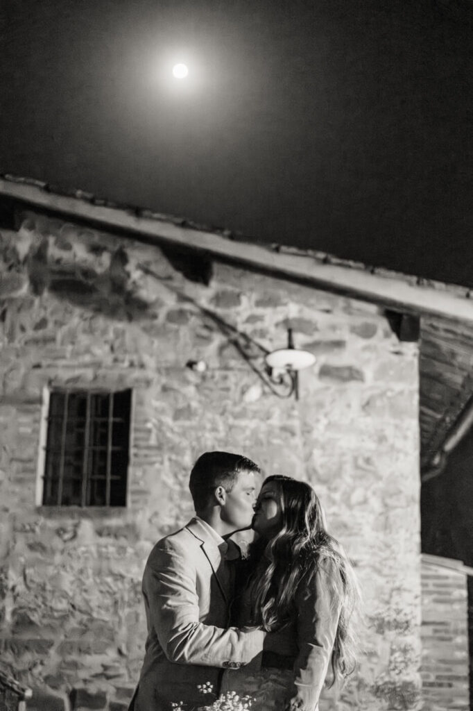 Bride and groom share a moonlit kiss at Villa Olimpia – A black and white image of the couple embracing outside the rustic stone villa, with the full moon glowing softly above them.