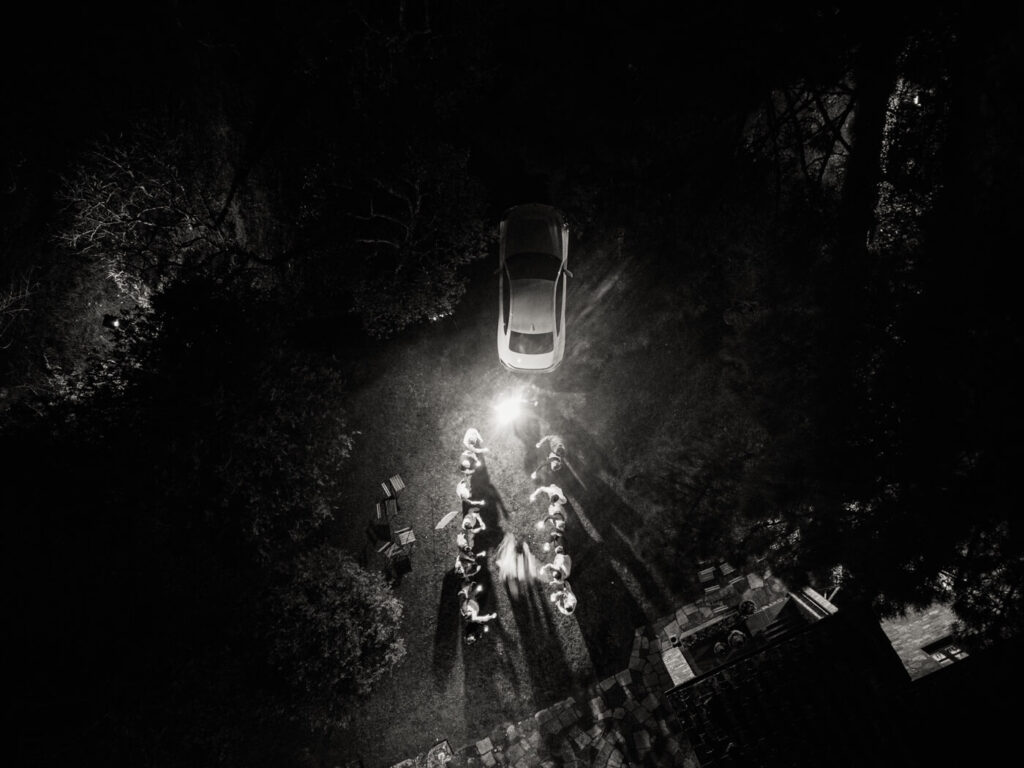 A cinematic wedding send-off beneath the stars – A black and white aerial image of the couple’s grand exit, as guests create a luminous pathway leading to their getaway car.