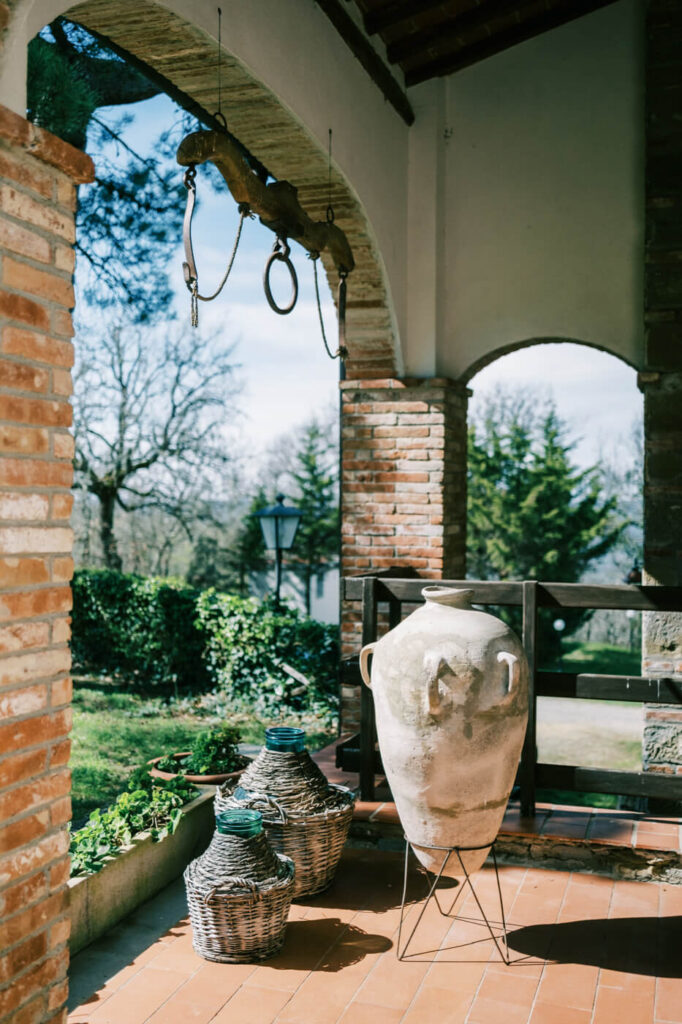 A sunlit Tuscan terrace at Villa Olimpia, featuring a large ceramic vase, woven baskets, and rustic brick archways.