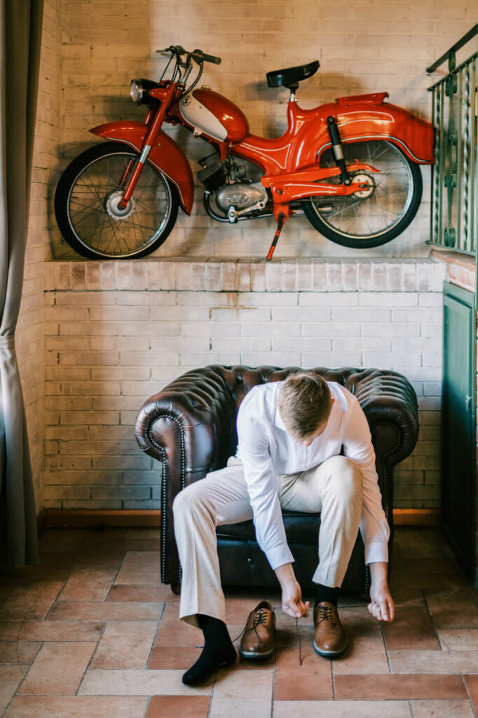 The groom prepares for his Tuscany villa wedding, sitting in a leather armchair while tying his shoes, with a vintage red motorcycle on the wall behind him.