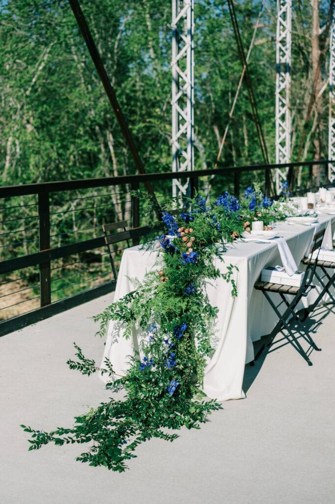 Elegant table setting at Finley Farms Bridge – A long reception table draped in white linen with cascading greenery and blue florals at Finley Farms Bridge in Missouri.