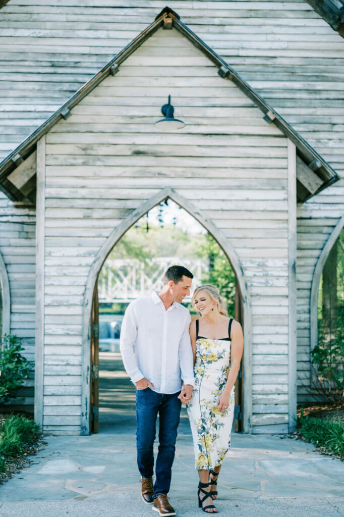 Couple Walking in Front of Rustic Chapel at Finley Farm Bridge Rehearsal Dinner – The bride and groom-to-be walk hand in hand, sharing a sweet moment in front of a charming wooden chapel at Finley Farm Bridge.