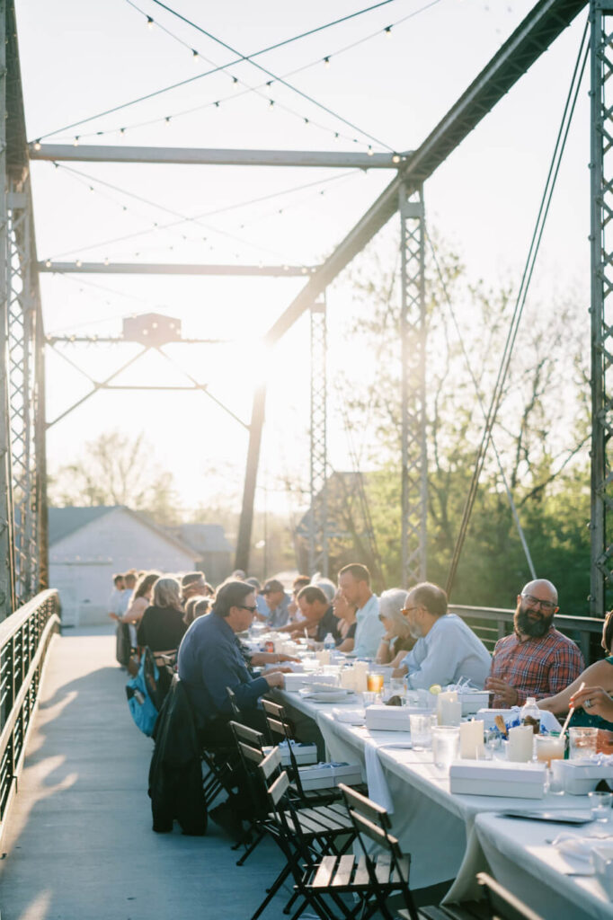 Guests Gathered for Rehearsal Dinner on Finley Farm Bridge Under String Lights – Loved ones share laughter and conversation at a beautifully set long table on Finley Farm Bridge, surrounded by nature.