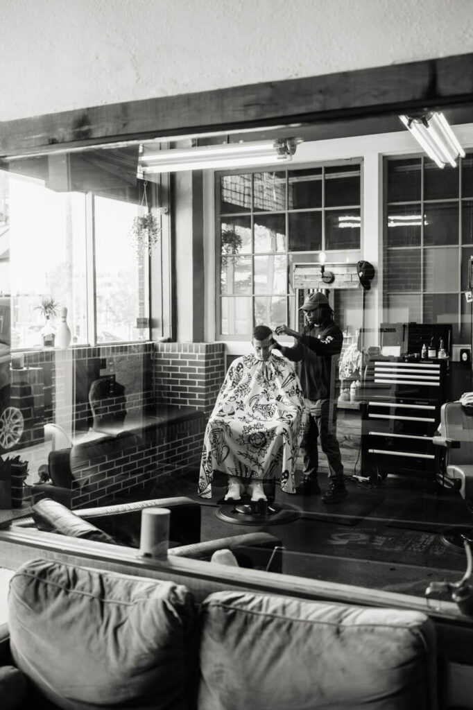 Groom's Preparation at the Barbershop – A black-and-white image of the groom getting a haircut at a modern barbershop, viewed through a glass window, capturing a quiet moment before the wedding.