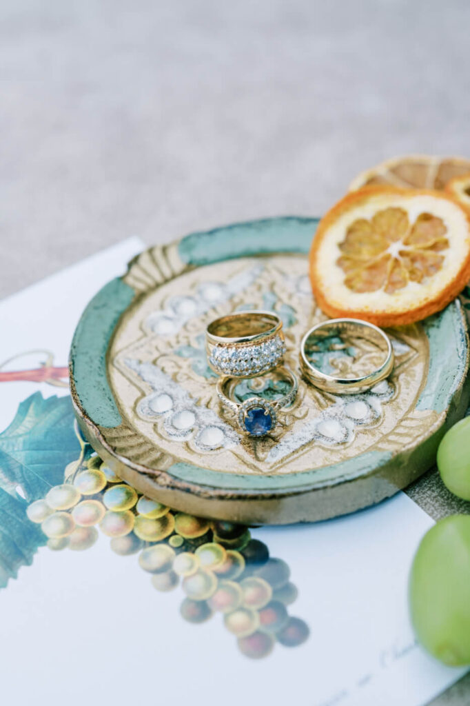 Exquisite Wedding Rings Display – A close-up of the wedding rings on an ornate ceramic dish, featuring a stunning sapphire engagement ring and gold wedding bands, with dried citrus slices as accents.