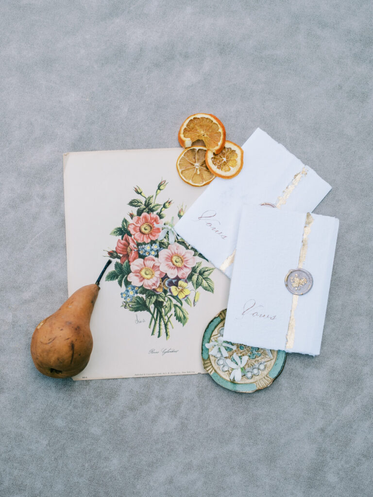 Romantic Wedding Vow Books – A flat-lay of handcrafted wedding vow books with wax seals, floral illustrations, and a pear, evoking a vintage-inspired aesthetic.