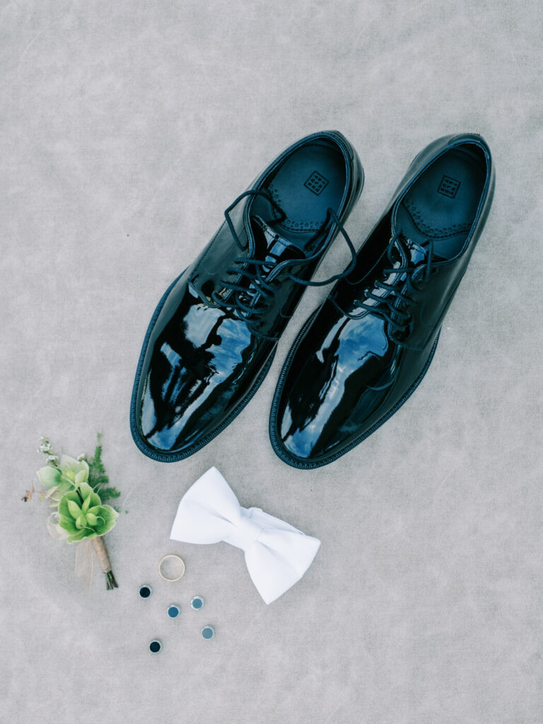 Groom's Wedding Accessories – A groom’s wedding details set, including polished black dress shoes, a crisp white bowtie, boutonniere, cufflinks, and wedding band, displayed on a neutral textured background.