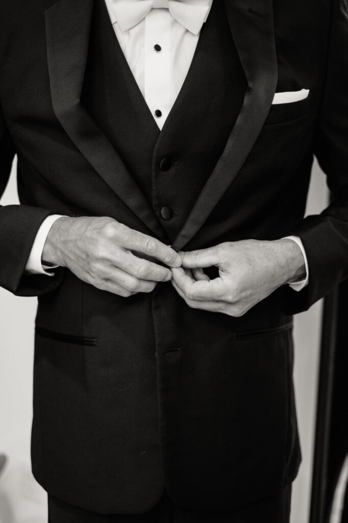 Groom Buttoning His Suit Jacket – A black-and-white image of the groom fastening the buttons on his classic black tuxedo, with a crisp white shirt and elegant bowtie.