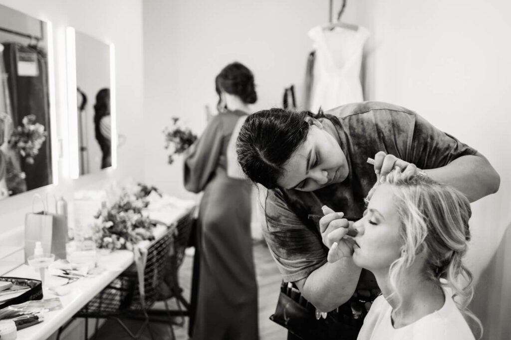 Bride Getting Her Makeup Done – A black-and-white documentary-style image of the bride getting her makeup done in a softly lit bridal suite, with a bridesmaid visible in the background.