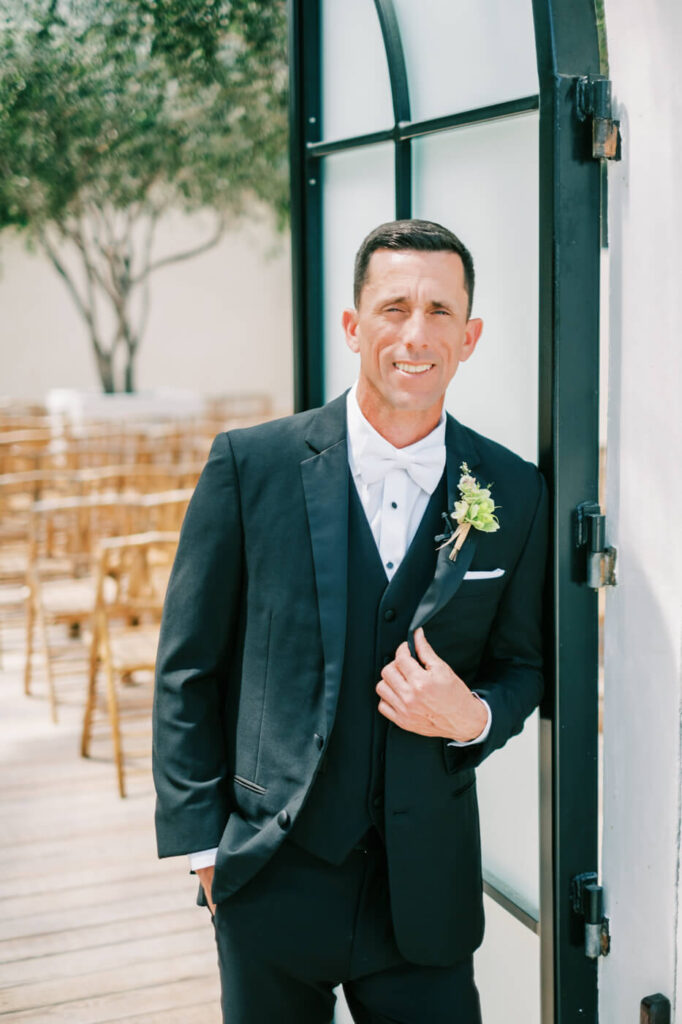 Groom Portrait at Two Rivers Greenhouse – A portrait of the groom in a black tuxedo with a white bowtie, leaning against a modern glass door, with wooden chairs set up in the background.