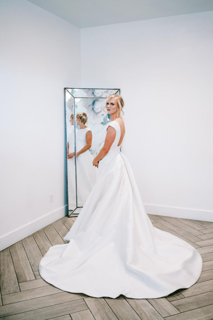 Bride’s Elegant Bridal Portrait – The bride standing gracefully in front of a full-length mirror, wearing a timeless white wedding gown with an open-back design and flowing train.