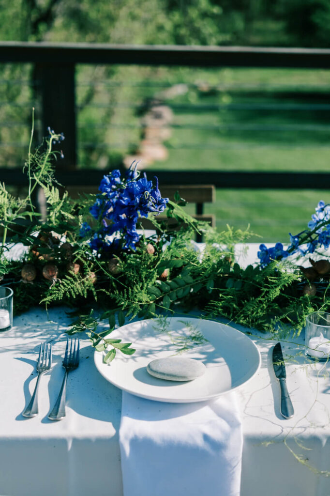 Place setting with floral garland at Finley Farms Bridge – A white plate with a smooth stone detail, surrounded by silverware and vibrant blue florals at the Finley Farms Bridge venue.