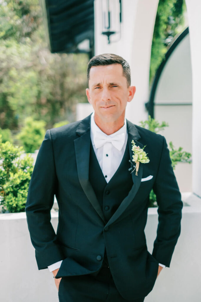 Groom Portrait at Two Rivers Greenhouse – A groom in a classic black tuxedo with a white bow tie stands confidently against a modern white archway, surrounded by lush greenery at Two Rivers Greenhouse.