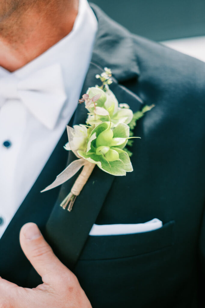 Groom’s Boutonniere Detail – A close-up of the groom’s lapel, showcasing a delicate green and ivory boutonniere wrapped in natural twine, complementing his elegant black suit.