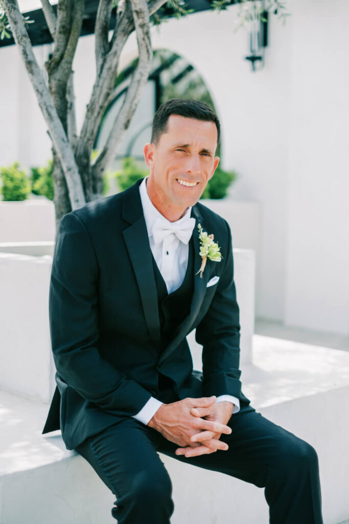 Groom Sitting Under an Olive Tree – The groom sits on a white stone bench under an olive tree, his hands folded as he smiles warmly, soaking in the moment before the ceremony.