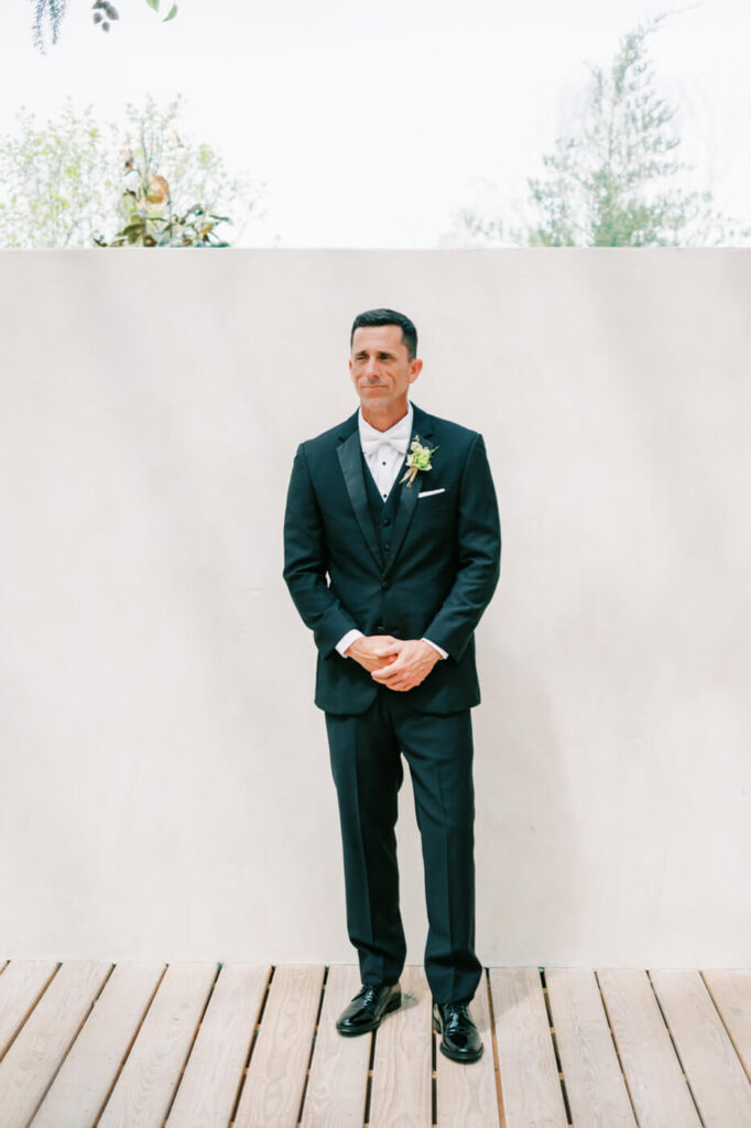 Groom Standing in Anticipation – A groom stands with hands folded in front of a minimalist white wall, dressed in a sharp black tuxedo with a fresh green boutonniere, waiting for his bride.