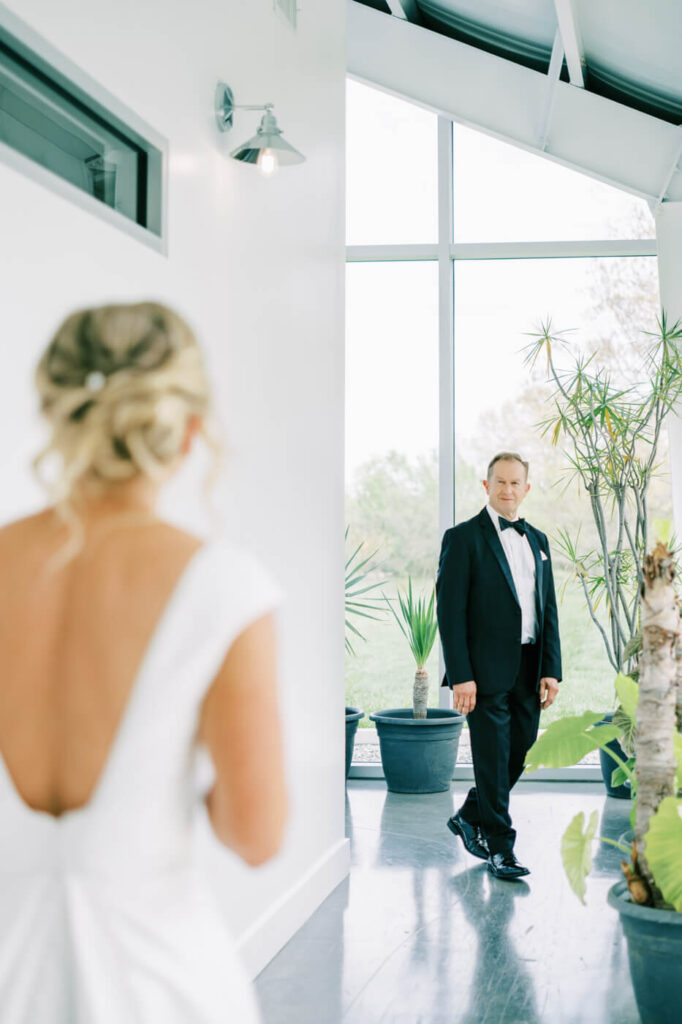 Father-Daughter First Look at Two Rivers Greenhouse – A bride in a white gown walks toward her father, who waits with an emotional expression inside the modern greenhouse setting.