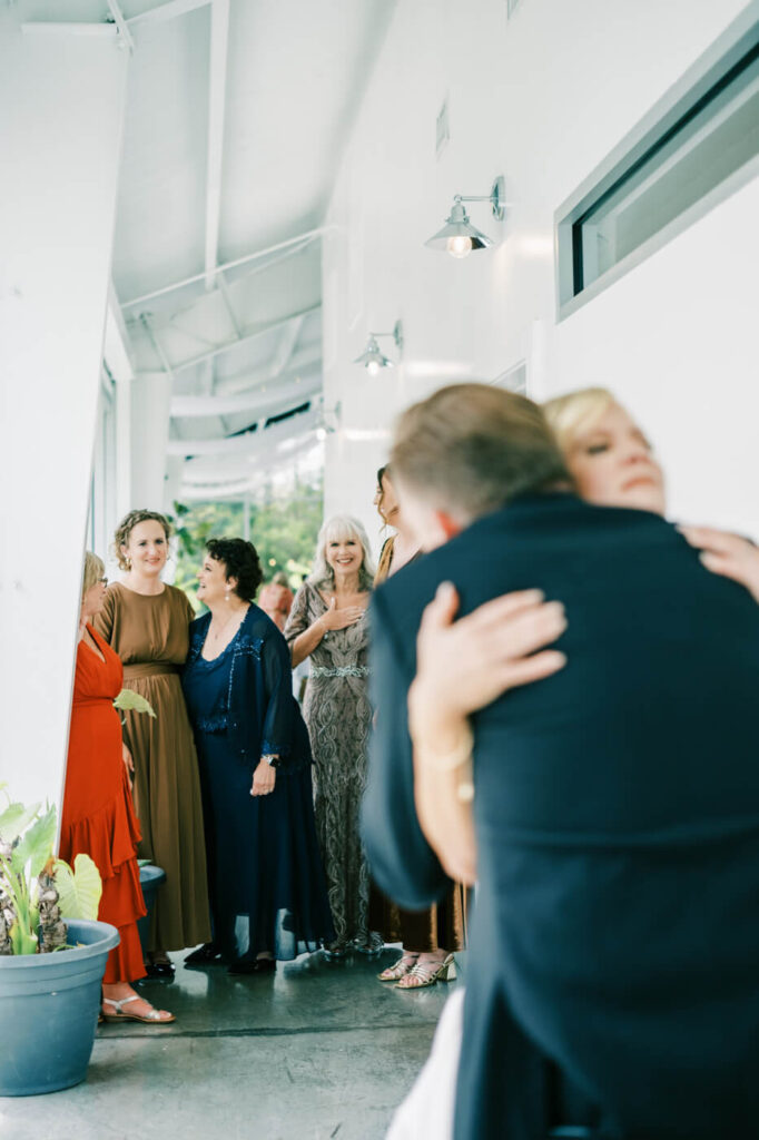 Emotional Embrace During First Look – The bride shares a heartfelt hug with a loved one as family members look on, their expressions filled with love and warmth in the airy, light-filled space.