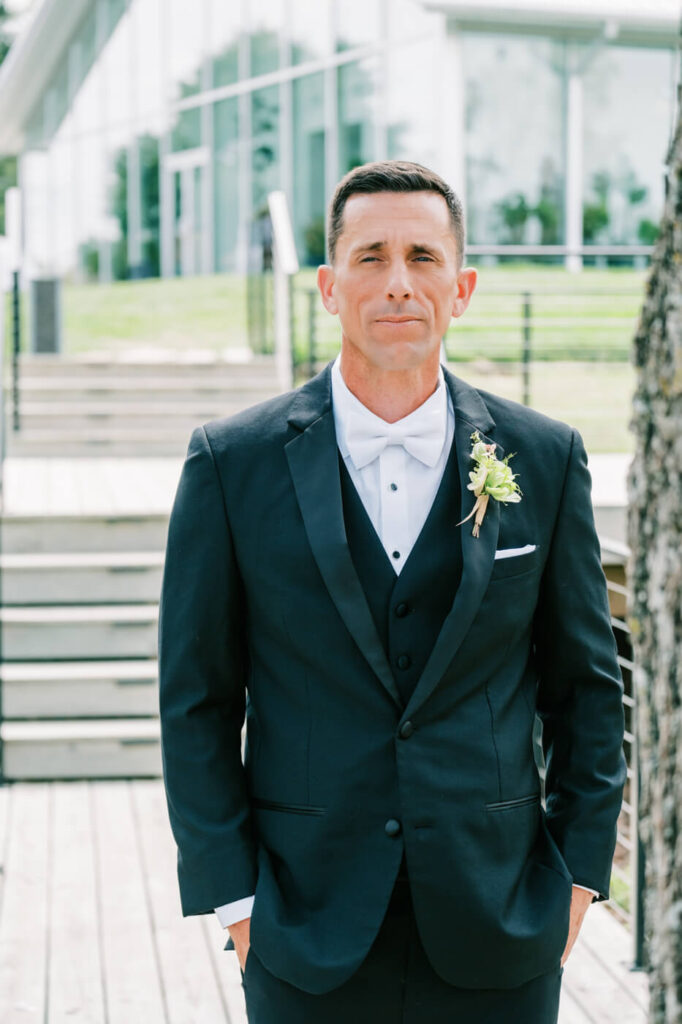 Groom Awaiting His Bride Outdoors – The groom stands outside Two Rivers Greenhouse, adjusting his stance as he prepares to see his bride, with a glass-walled venue and natural landscape in the background.