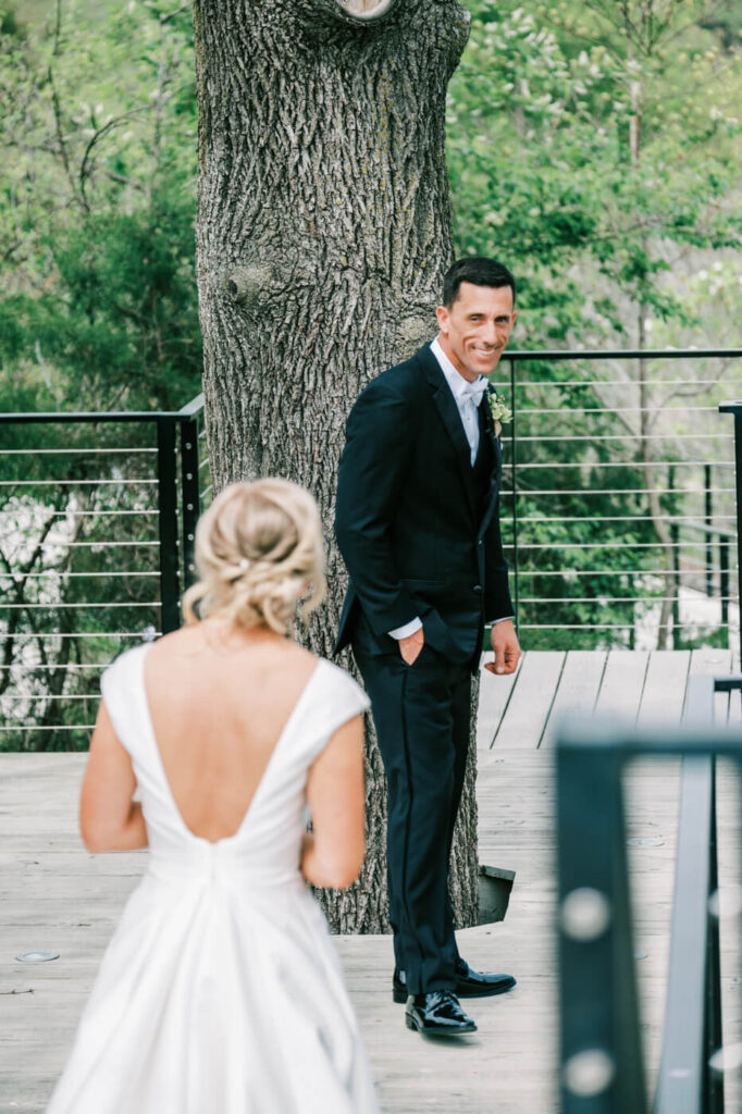 First Look on the Outdoor Deck – The groom, smiling, turns to see his bride for the first time, standing near a tree-lined deck, surrounded by greenery and rustic railings.