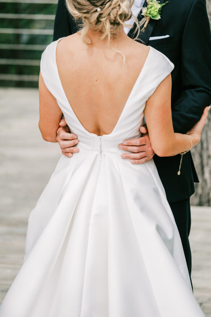 Bride and Groom’s Tender Embrace – The groom wraps his arms around the bride’s waist as she stands in her timeless white gown, her back to the camera, capturing the intimacy of the moment.