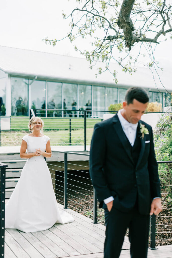 Bride Walking Toward Groom for First Look – The bride, beaming with excitement, approaches her groom, who stands with his back turned on a scenic outdoor deck, with the modern greenhouse venue reflected in the glass windows behind them.