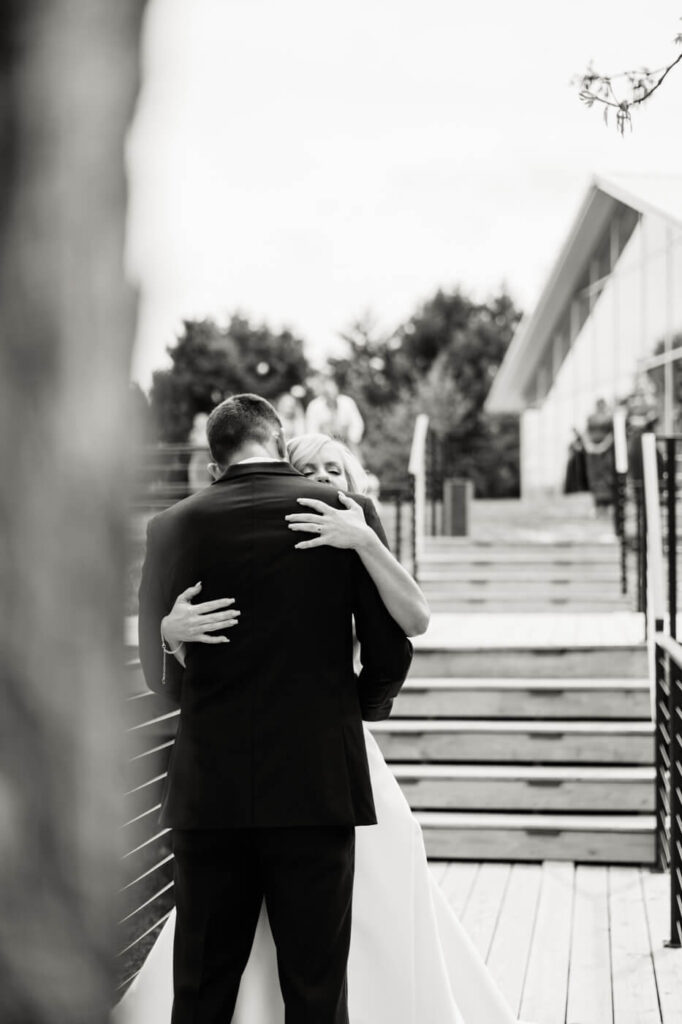 Bride and groom first look embrace at The Greenhouse Two Rivers – A black and white image of a bride and groom holding each other tightly on a wooden deck at The Greenhouse Two Rivers, with soft-focus greenery and wedding guests in the background.