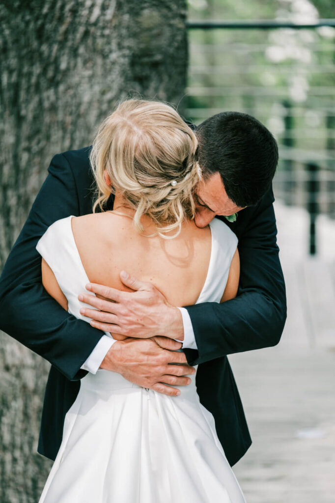 Emotional first look hug at The Greenhouse Two Rivers wedding – A groom in a black tuxedo embraces his bride with deep emotion, his head tucked into her shoulder, set against the backdrop of a large tree at The Greenhouse Two Rivers.