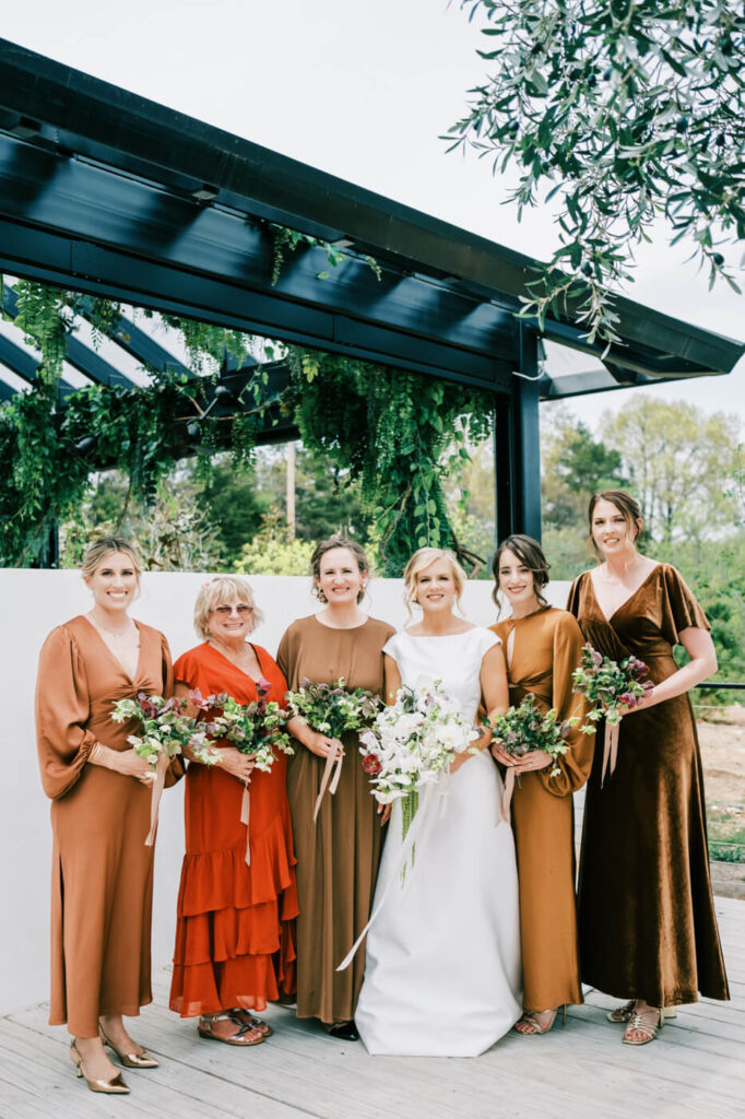 Bride with bridesmaids in earth-toned dresses at The Greenhouse Two Rivers – The bride stands with her bridesmaids dressed in warm rust, brown, and gold tones, holding bouquets of greenery and florals beneath a modern black pergola draped in vines at The Greenhouse Two Rivers.