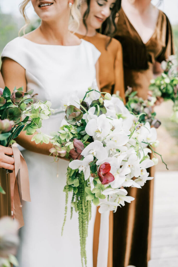 Bride’s cascading white orchid bouquet at The Greenhouse Two Rivers wedding – The bride holds an elegant cascading bouquet of white orchids, burgundy blooms, and trailing greenery, surrounded by bridesmaids in earth-toned dresses at The Greenhouse Two Rivers.