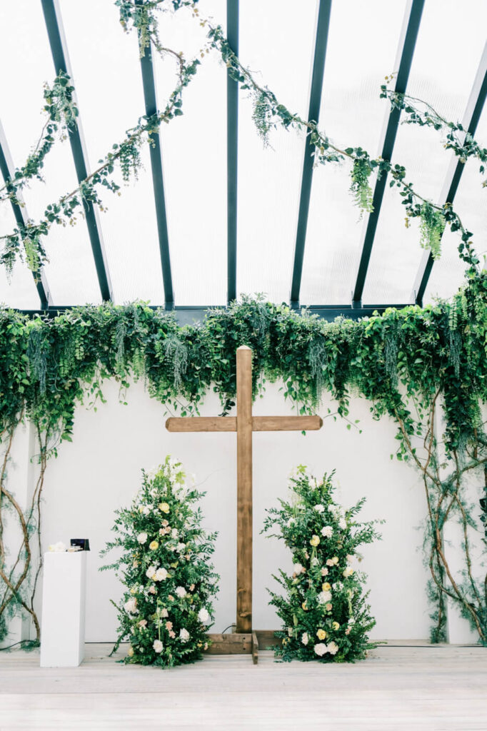 Ceremony setup at The Greenhouse Two Rivers with wooden cross and lush greenery – A wedding ceremony altar featuring a wooden cross, flanked by floral arrangements, with greenery draping from the ceiling in a greenhouse-inspired venue at The Greenhouse Two Rivers.