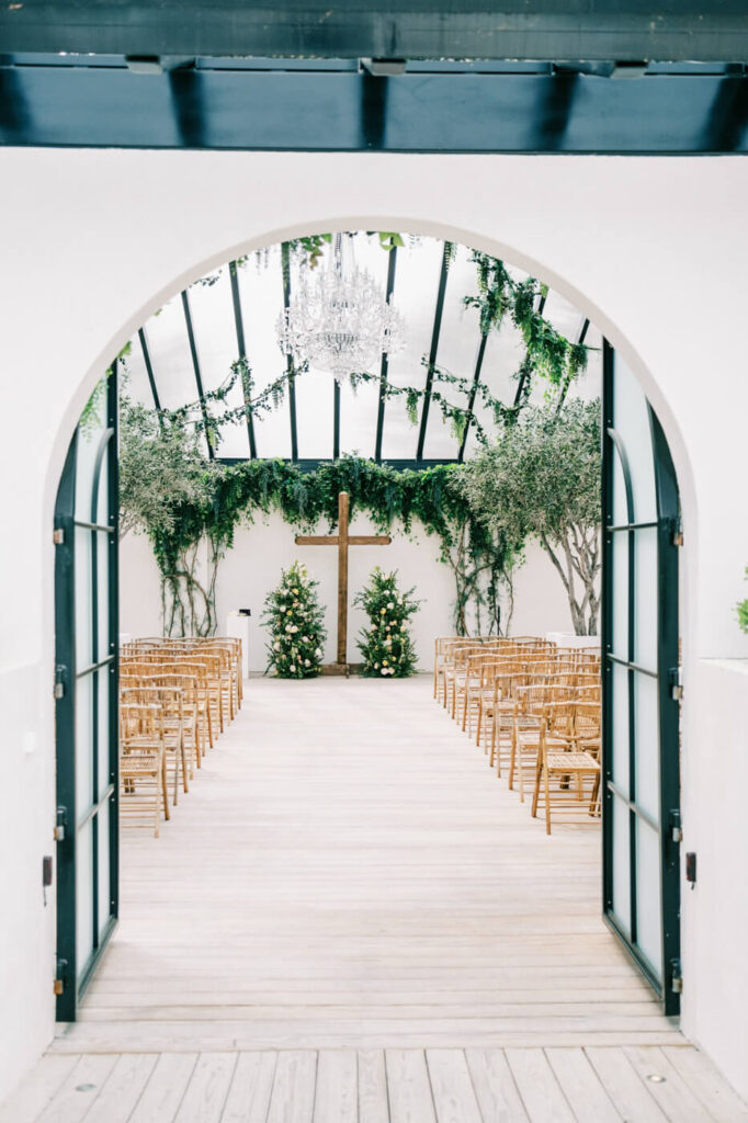 Wedding ceremony view through arched doorway at The Greenhouse Two Rivers – A bright ceremony space framed by an arched entryway at The Greenhouse Two Rivers, showing wooden chairs, lush greenery, and a floral-adorned wooden cross as the altar.