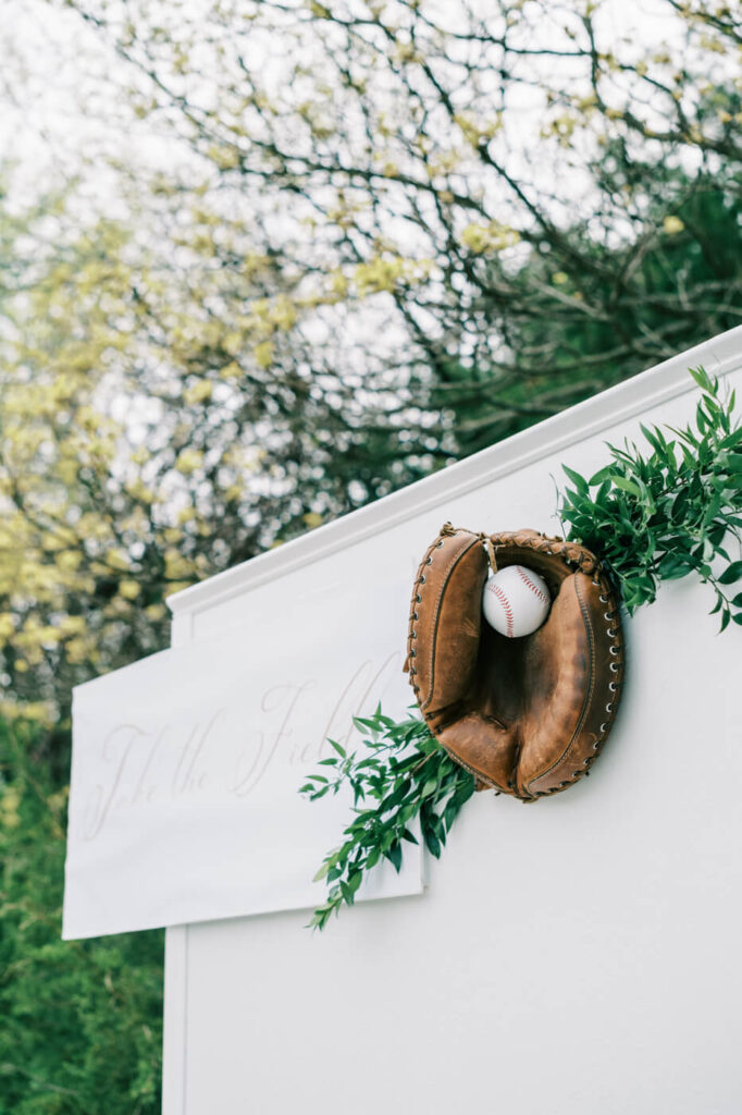 Close-up of baseball glove and ball on seating chart at The Greenhouse Two Rivers – A vintage baseball glove with a baseball mounted on a white seating chart wall, decorated with greenery and elegant calligraphy at The Greenhouse Two Rivers.